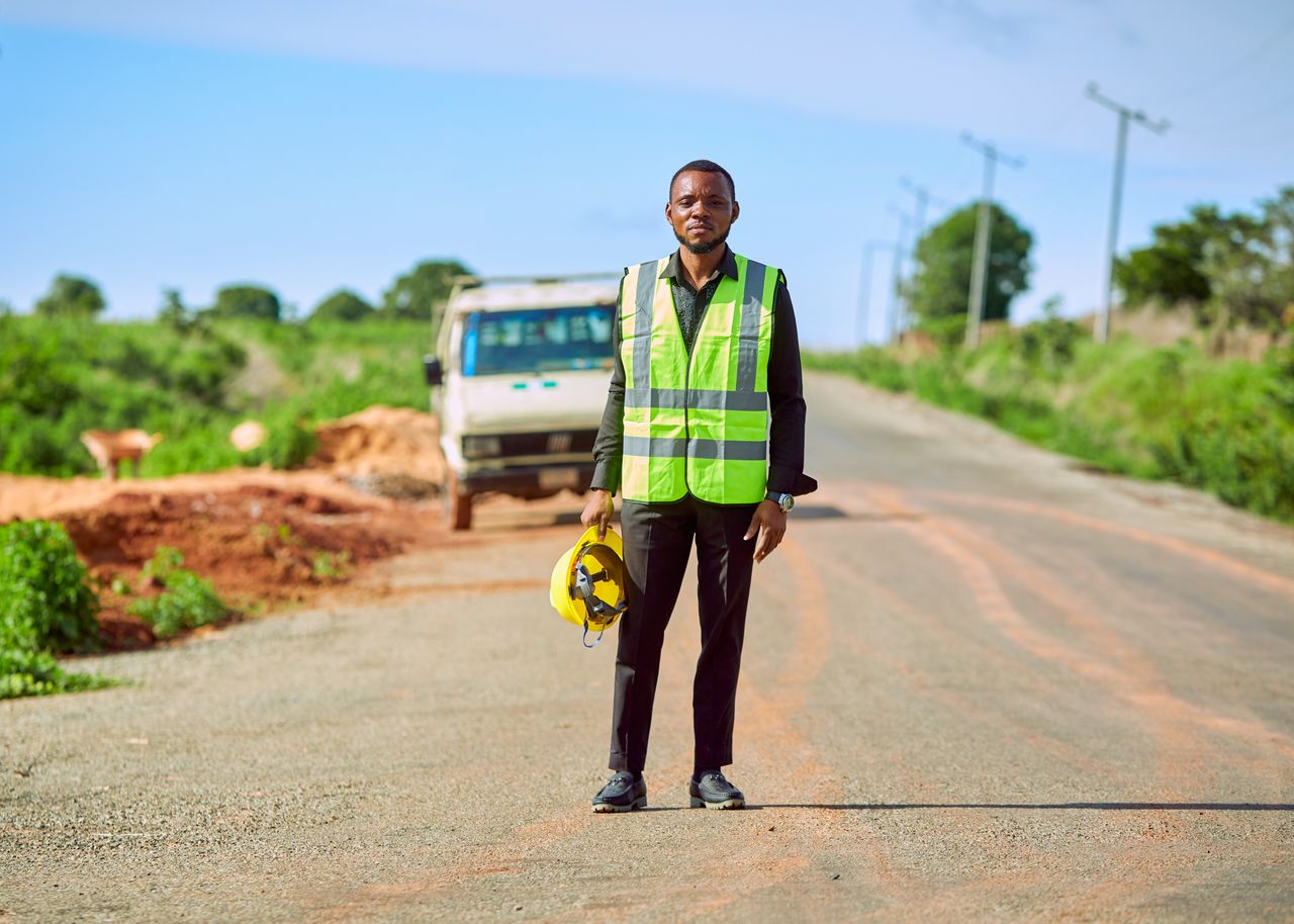Crafting Captivating Headlines: Your awesome post title goes here A construction worker in a safety vest stands on a rural road in Lafia, Nigeria, holding a helmet.